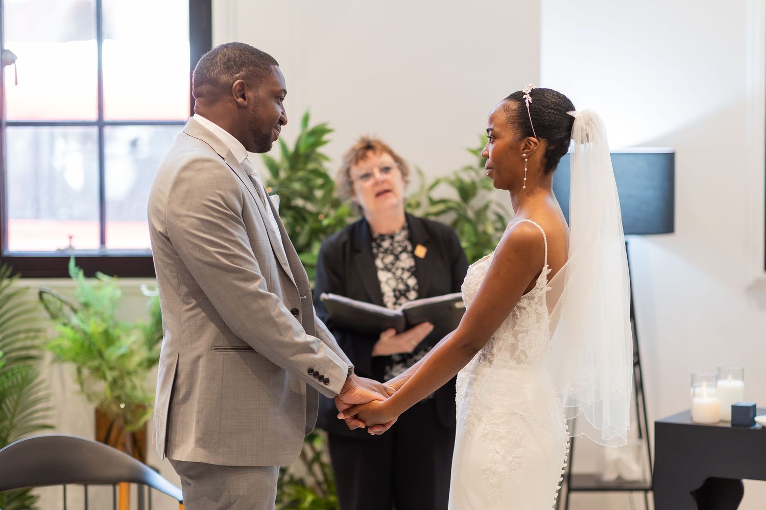 A bride and groom hold hands during their wedding ceremony while an officiant reads vows in an elegant indoor venue.