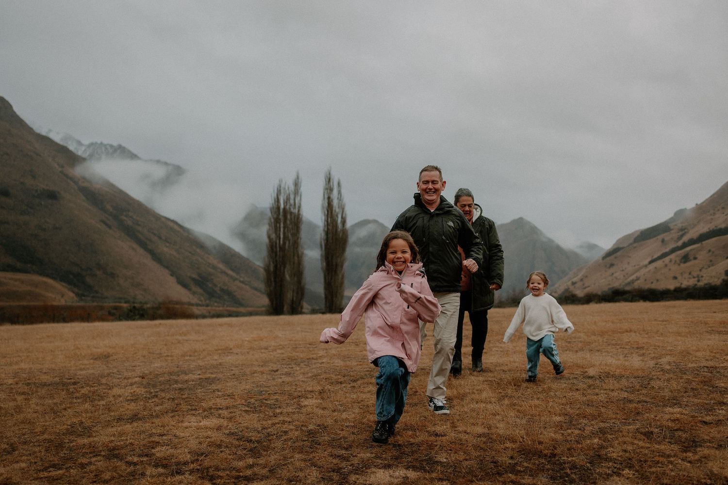 Family running and laughing in a misty mountain valley with snow-capped peaks and golden grasslands.