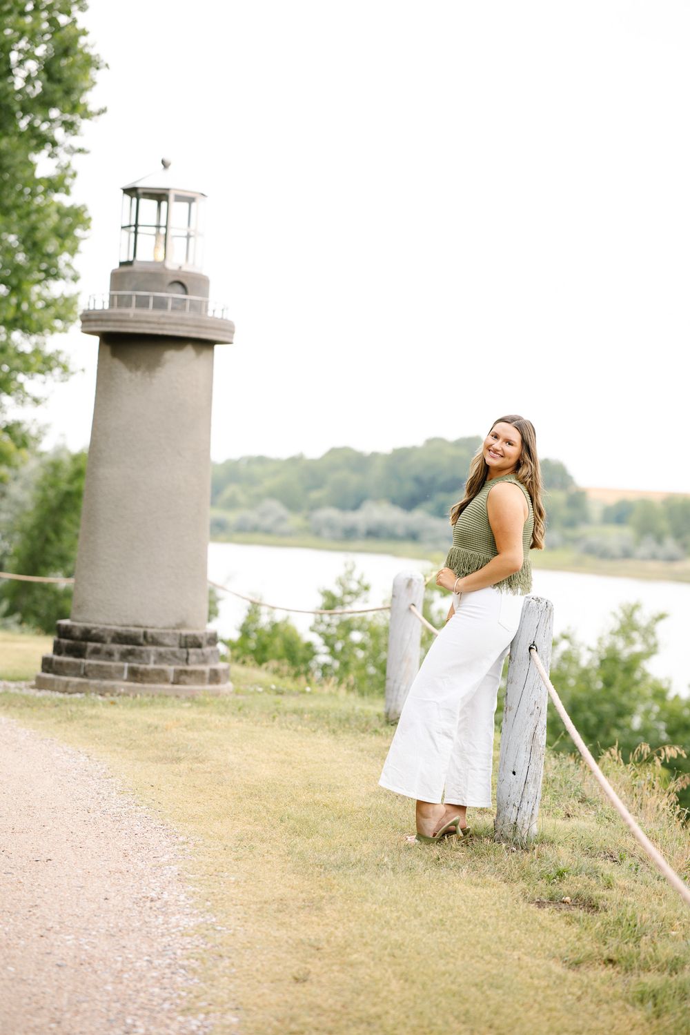 Young woman in white skirt and green top smiling near a small lighthouse by a serene lakeside path.