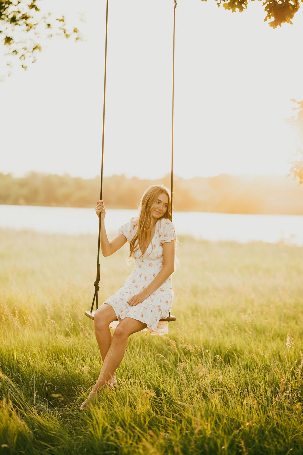 Young woman in floral dress sitting on rope swing at golden sunset near lake surrounded by lush green grass.