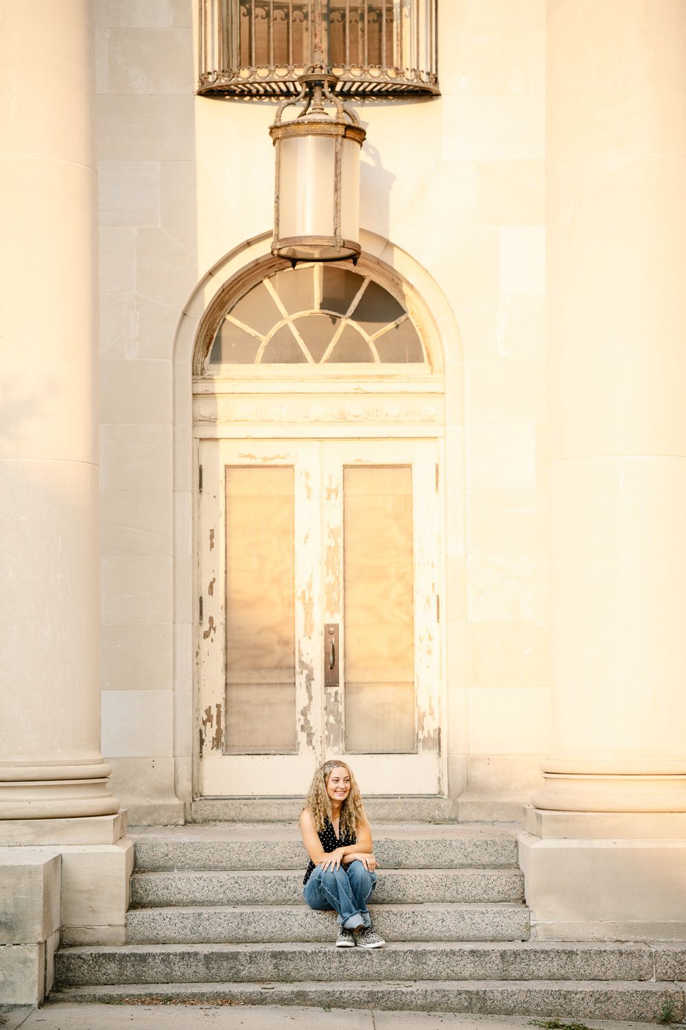 Young woman with curly hair sitting on stone steps in front of an ornate arched doorway of a classic building.