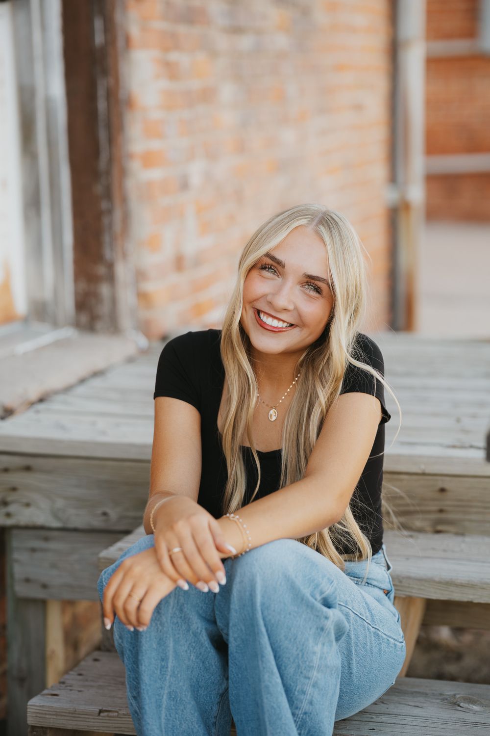 Smiling blonde woman in black top and jeans sitting on wooden steps in front of a brick wall.
