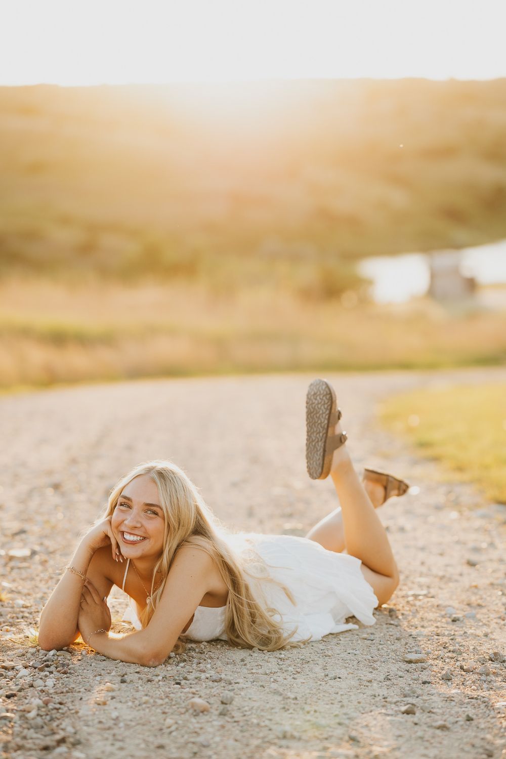 Smiling blonde woman in white dress lying on a dirt path during golden hour sunset in a rural outdoor setting.