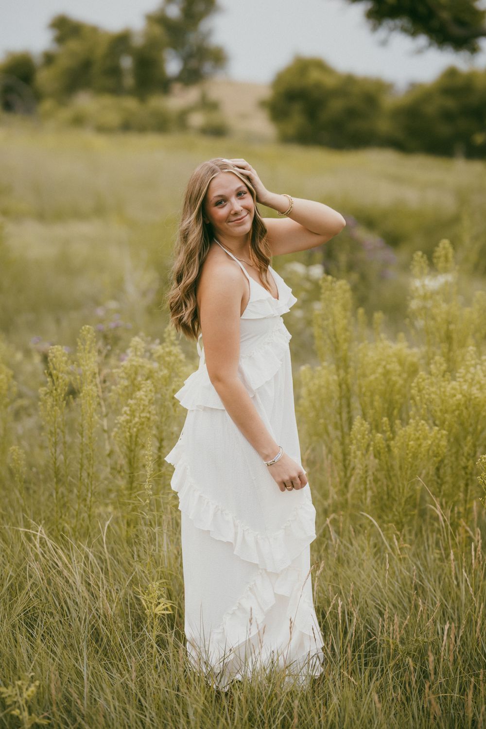 Young woman in white ruffled maxi dress standing in a lush green wildflower field, smiling softly.