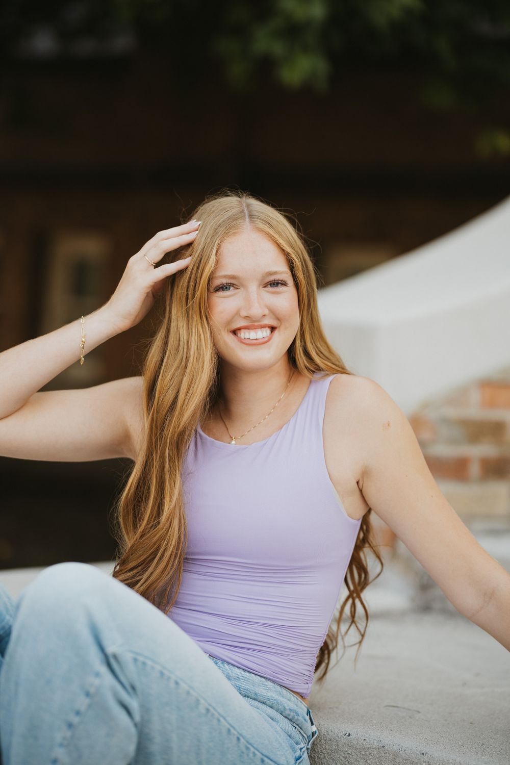Smiling young woman with long auburn hair wearing a lavender tank top and jeans, sitting outdoors in warm sunlight.