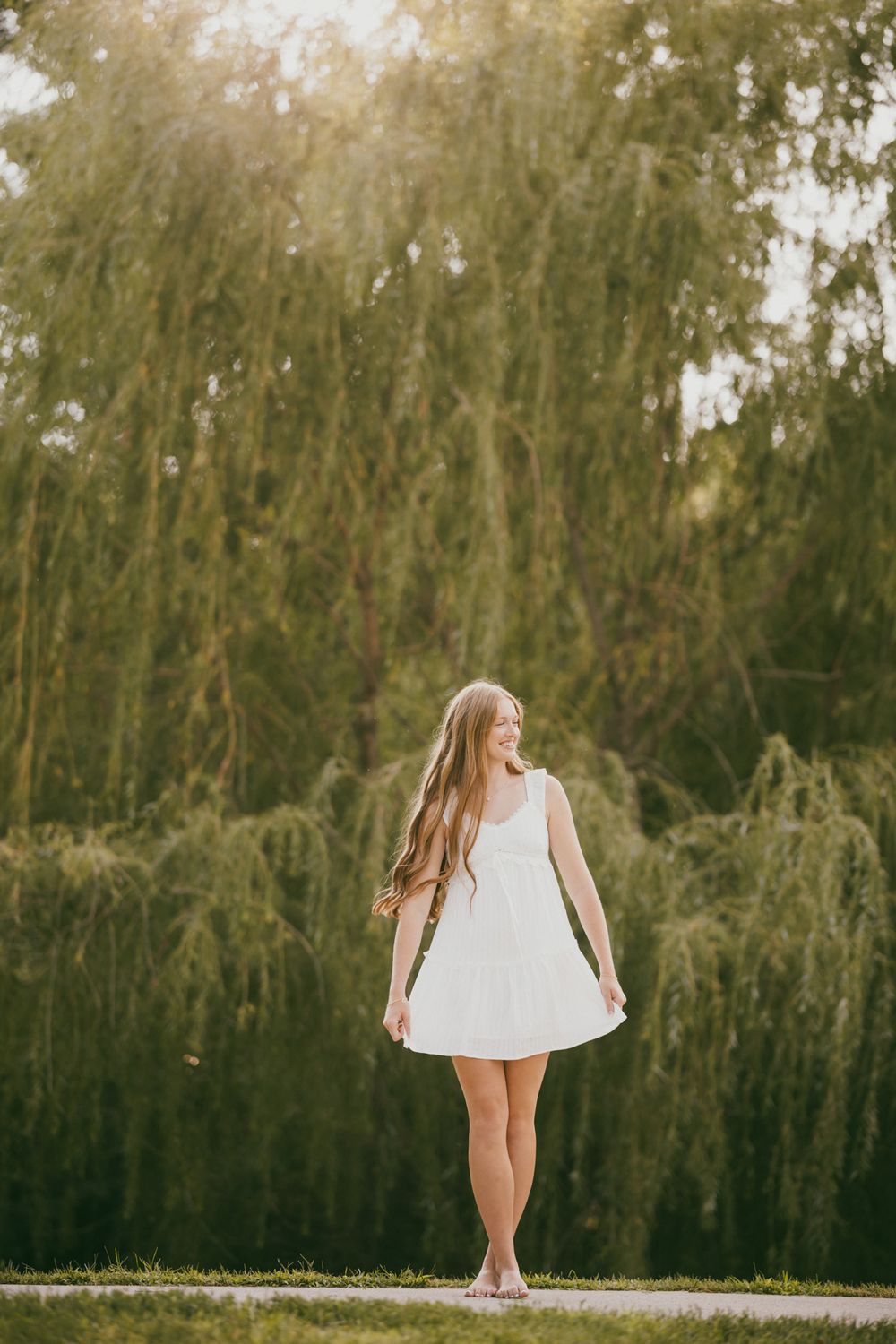Young woman in white dress standing barefoot in front of lush weeping willow trees in golden sunlight.