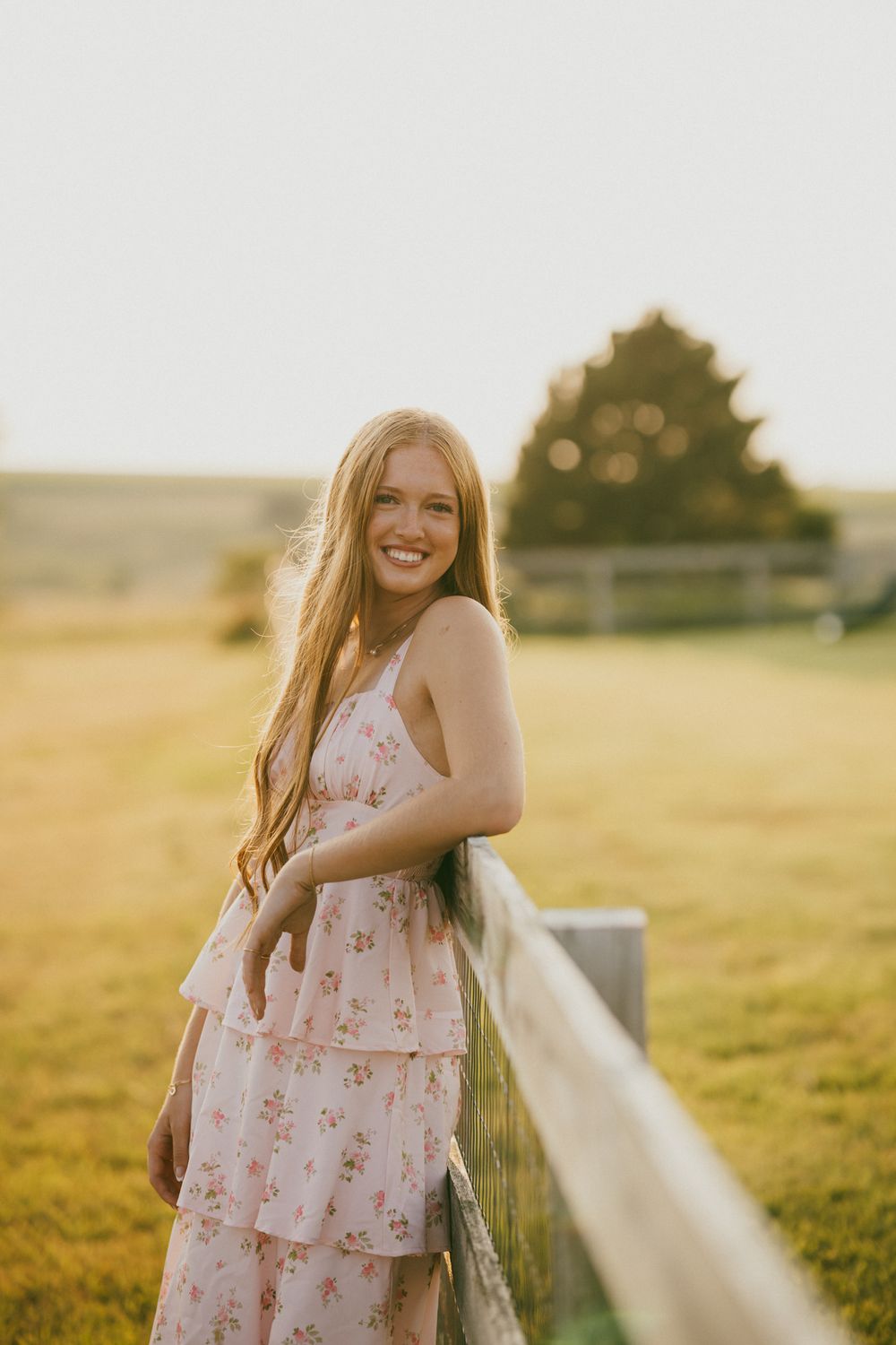 Young woman in floral pink dress leaning on wooden fence in golden hour sunlight in a green field.