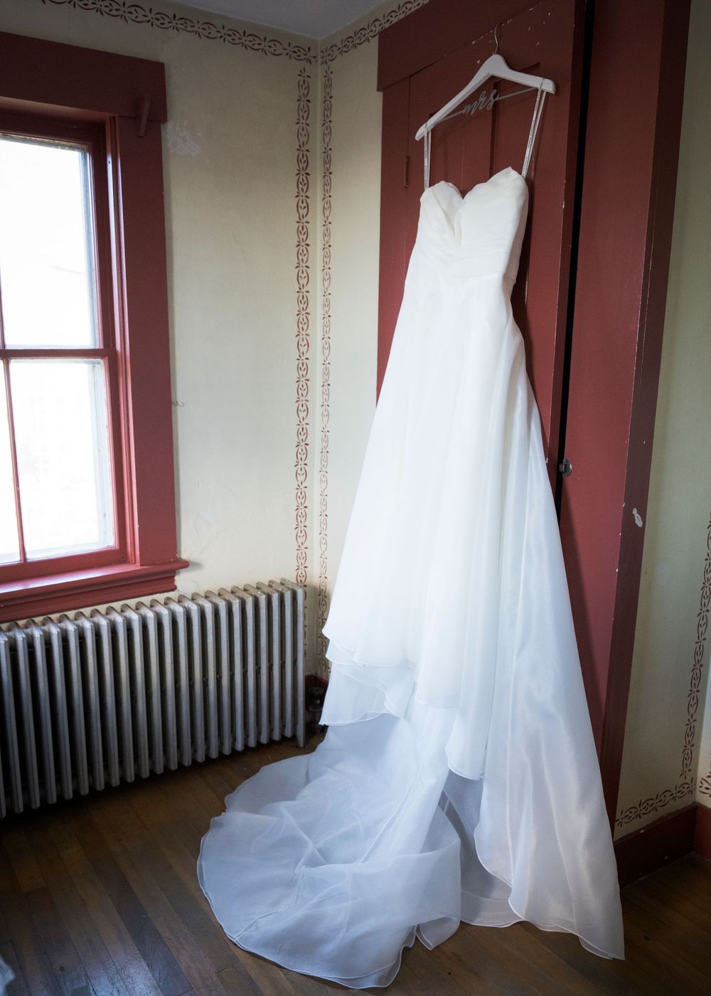 White strapless wedding gown with long train hanging on a hanger in a vintage room with red trim windows.