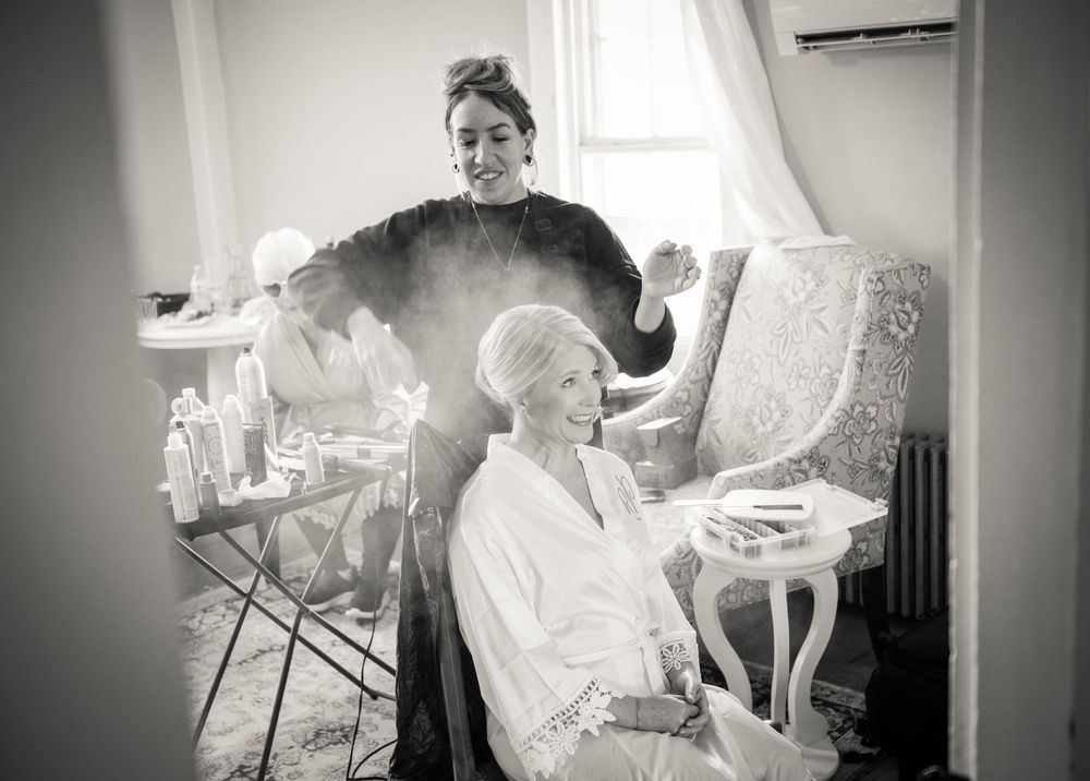 Black and white photo of bride getting hair and makeup done by stylists before her wedding.