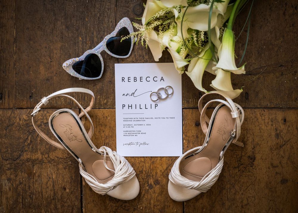 Wedding flat lay featuring invitation for Rebecca and Phillip, bridal heels, rings, sunglasses, and flowers.