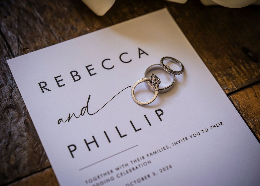 Wedding invitation for Rebecca and Phillip with elegant rings resting on white card on rustic wood surface.