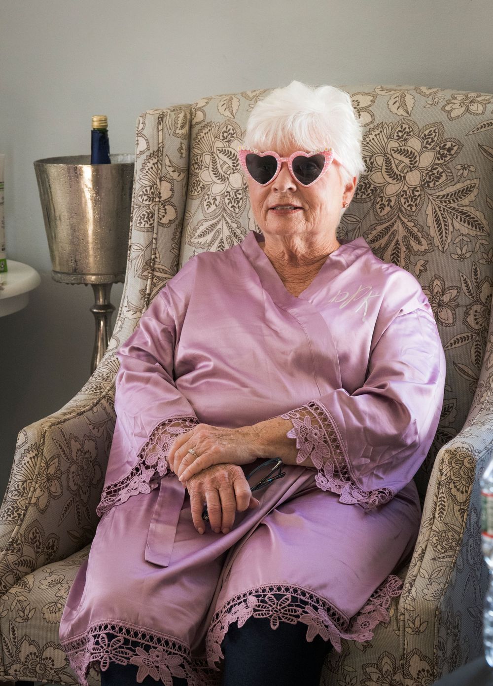 Elderly woman in pink satin robe and heart-shaped sunglasses seated in floral armchair.