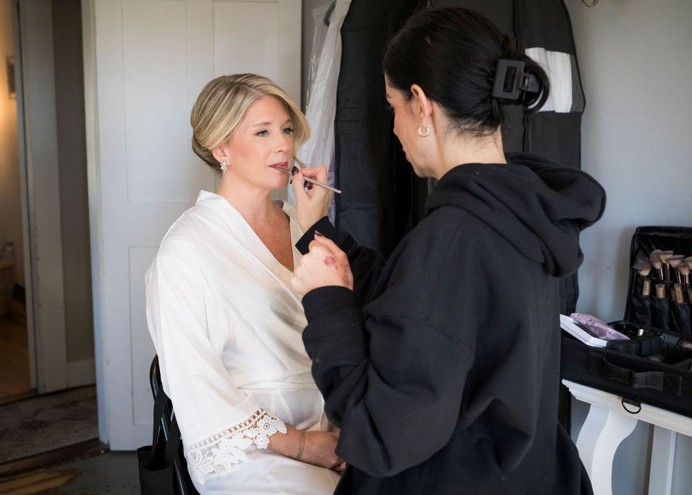 Makeup artist applying lipstick to blonde bride in white robe during wedding preparation.