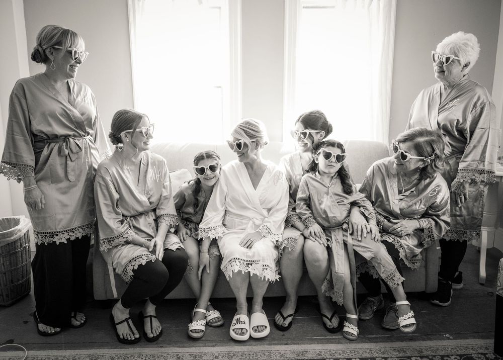 Bride and bridesmaids wearing robes and fun sunglasses pose together in a bright room during wedding preparations.