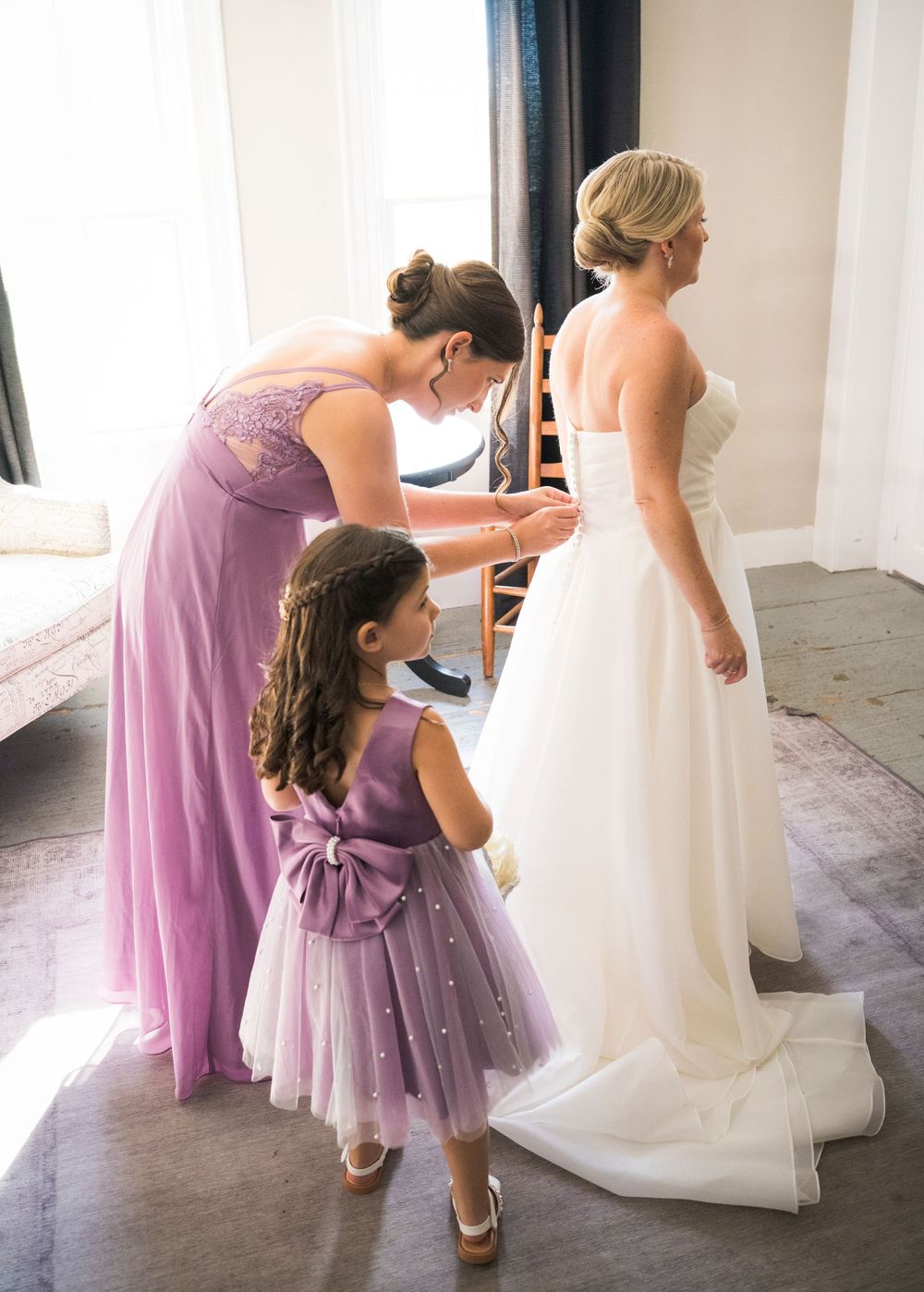 Bridesmaid in purple gown and flower girl helping bride button her white wedding dress before ceremony.