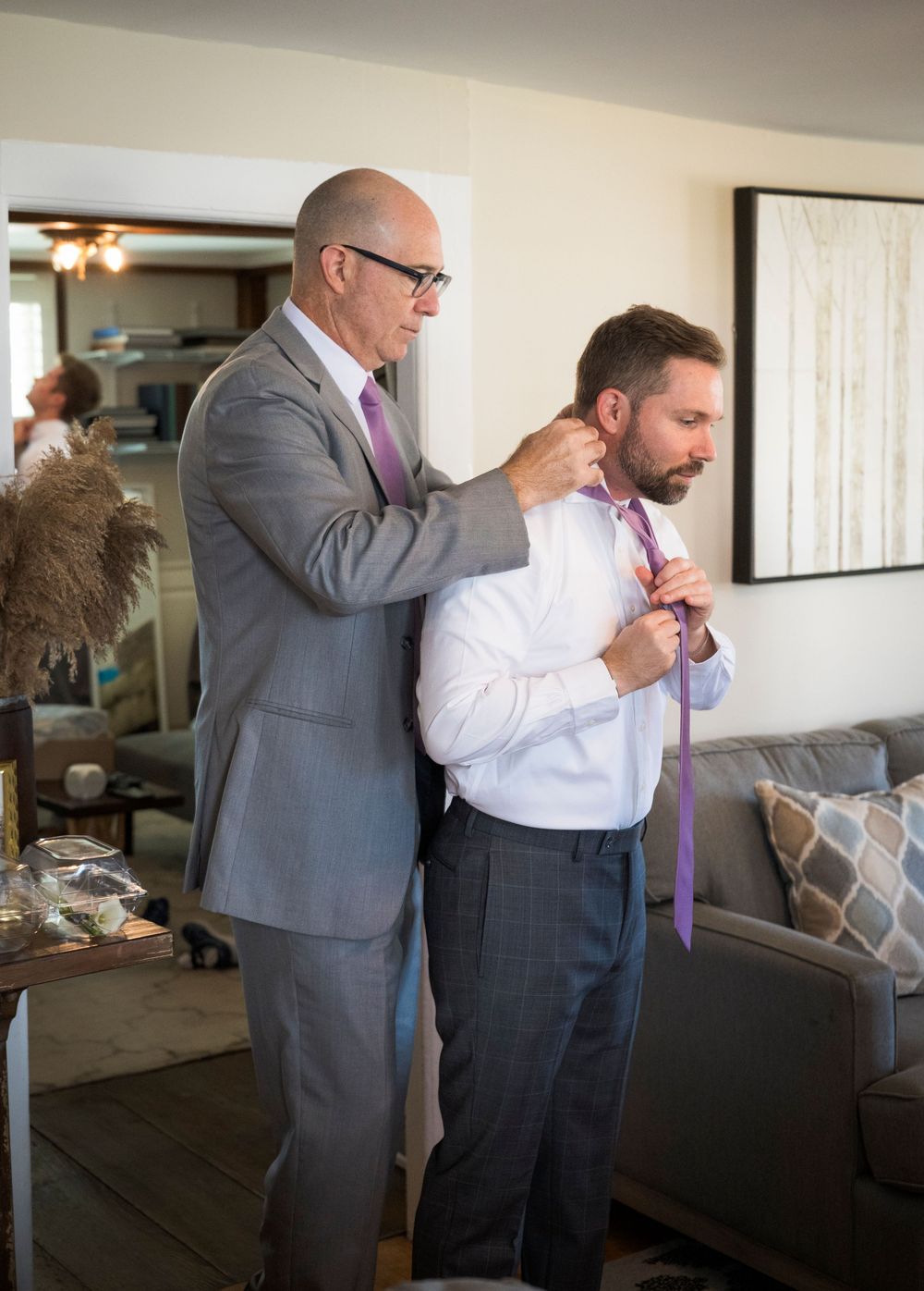 Older man in gray suit helping younger man adjust purple tie indoors.