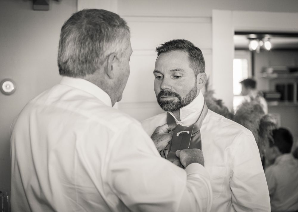 Black and white photo of a man helping another man adjust his tie before a wedding ceremony.