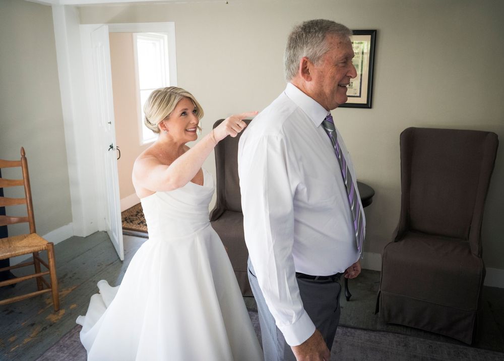 Bride in white gown helping older man with his collar inside a softly lit room before the wedding.