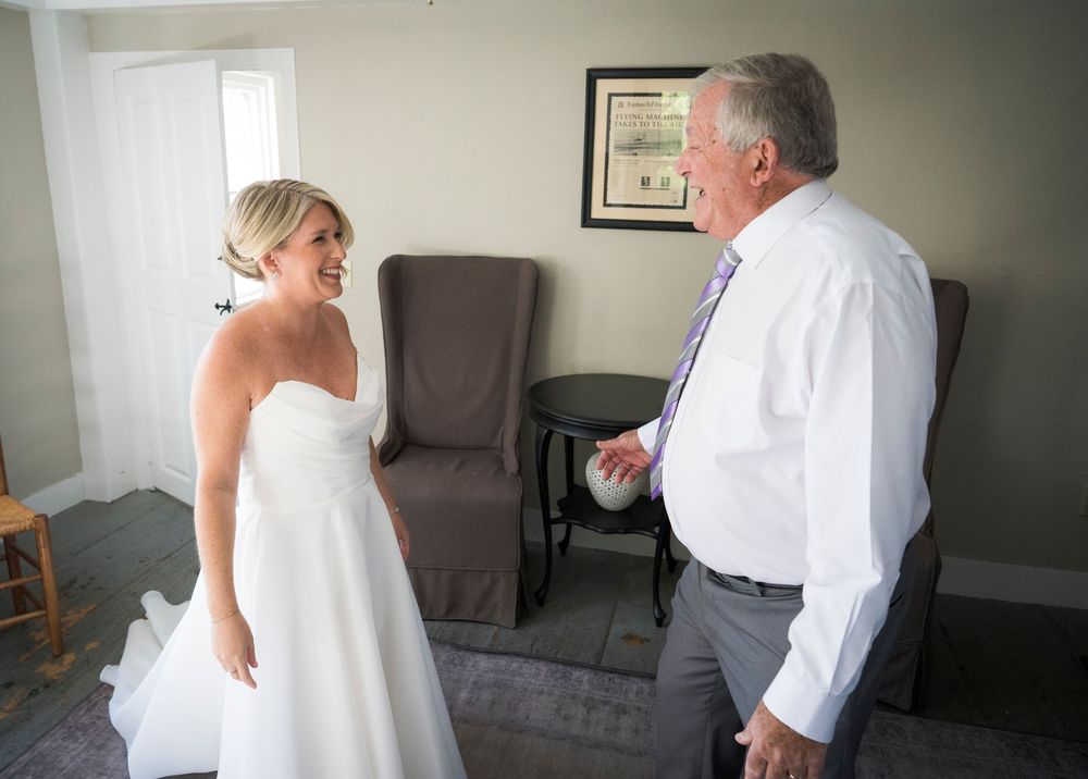 Bride in white strapless gown sharing first look moment with father in hotel room.