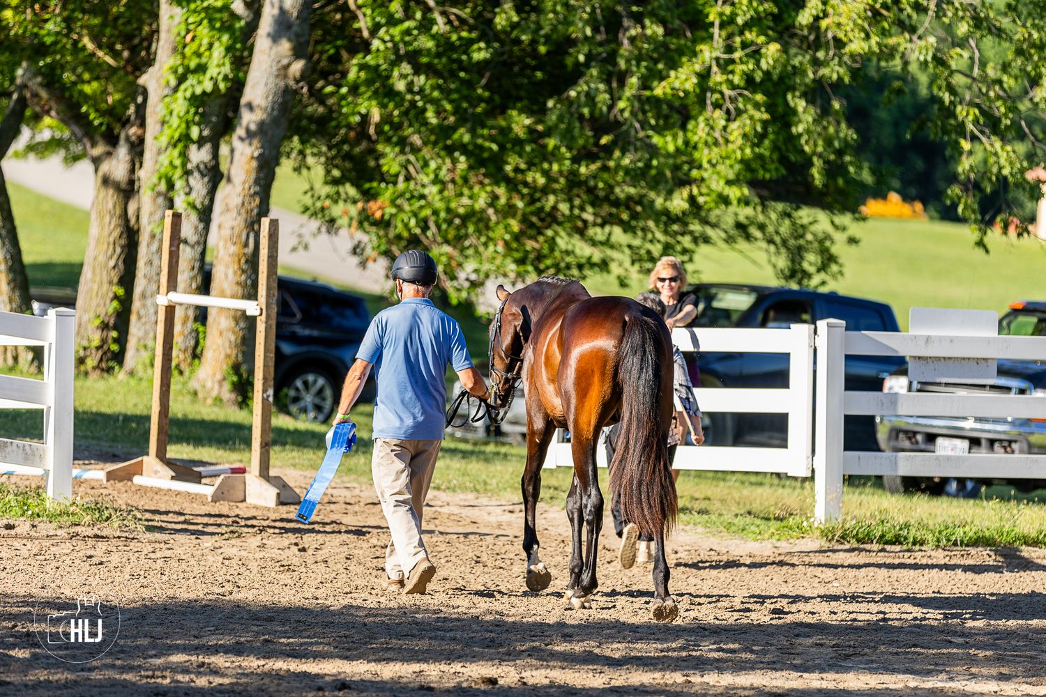 Two handlers walking a bay horse along a white-fenced paddock path surrounded by lush green trees.
