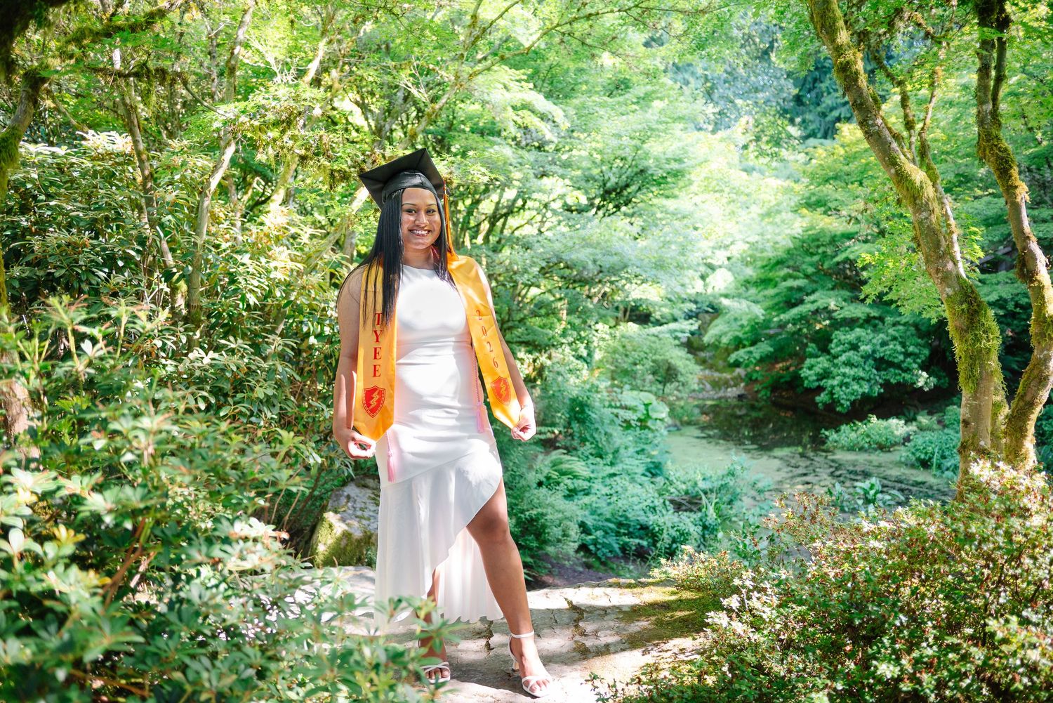 Graduate in cap and gold stole standing on a stone surrounded by the green landscape of the Yao Garden