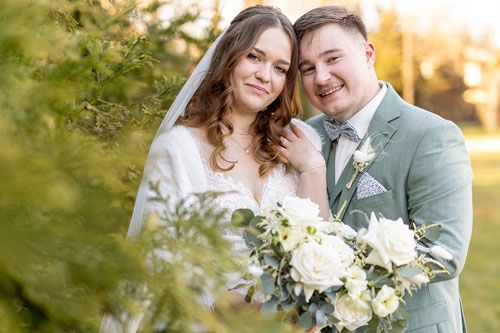 Bride and groom pose together outdoors at golden hour, she holds white floral bouquet, he wears sage suit with boutonniere.