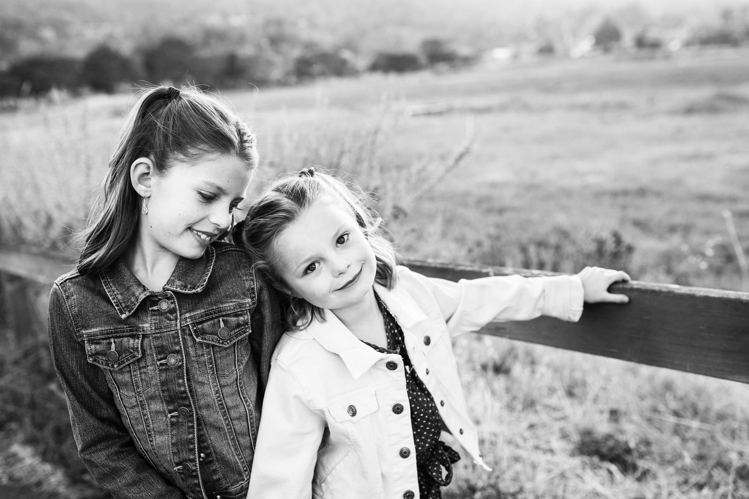 Two young girls pose by a wooden fence in a black and white outdoor portrait photo.