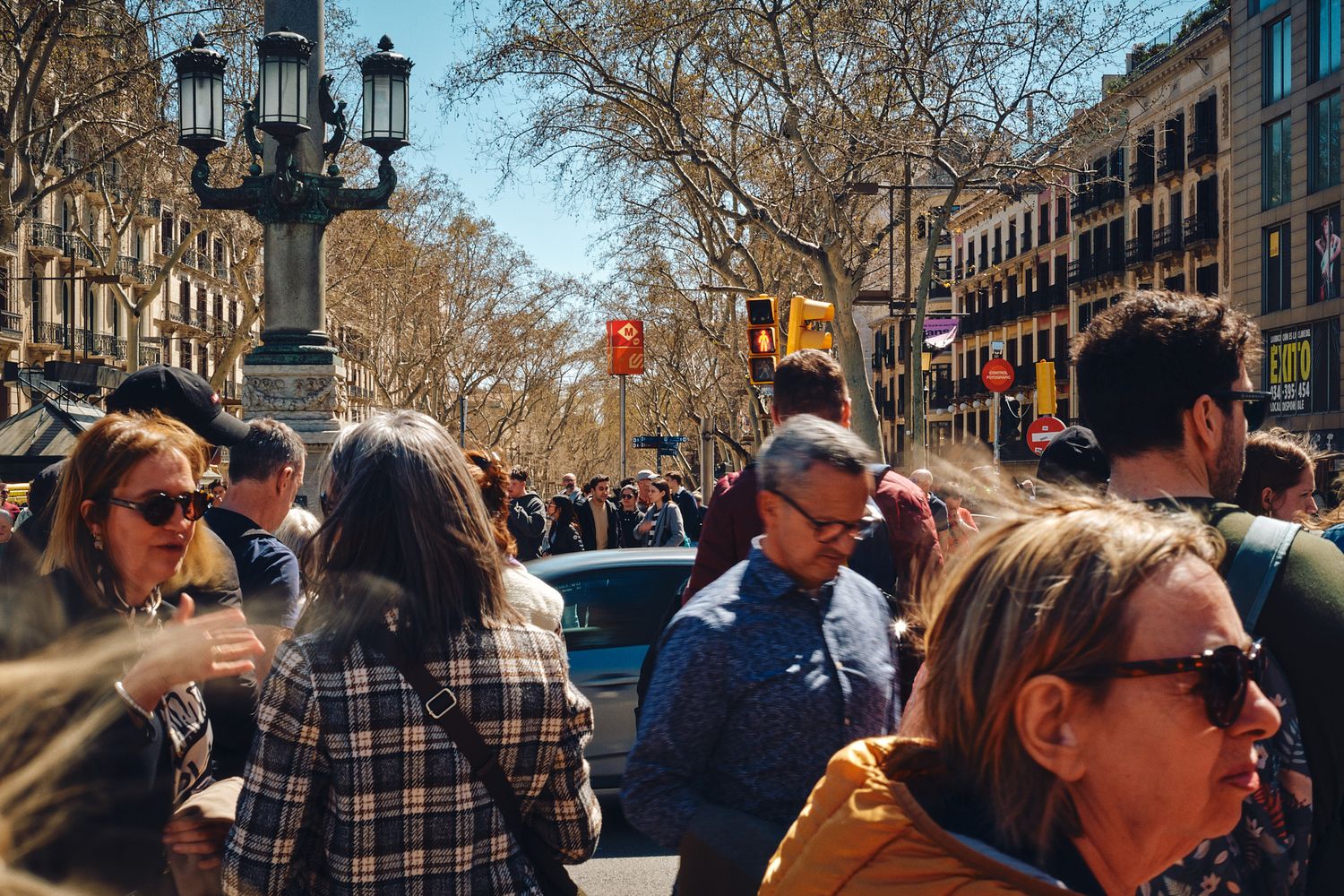 Crowded street scene on La Rambla, Barcelona, with tourists and locals walking under bare trees on a sunny day.