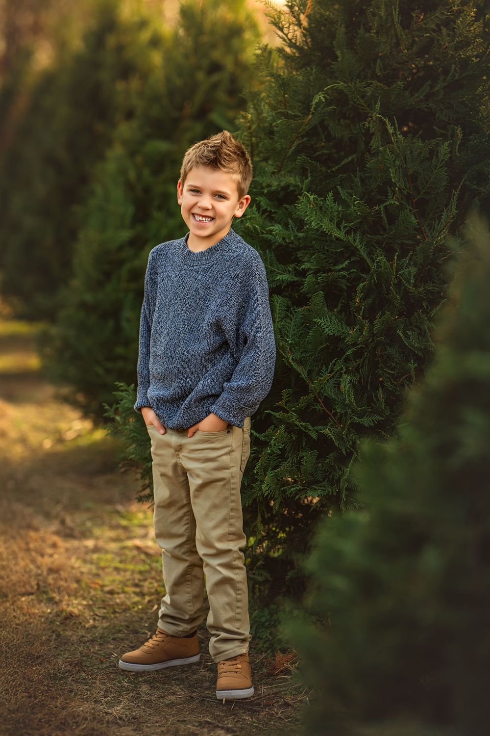 A smiling boy in a blue sweater and khaki pants stands among tall green evergreen trees outdoors.