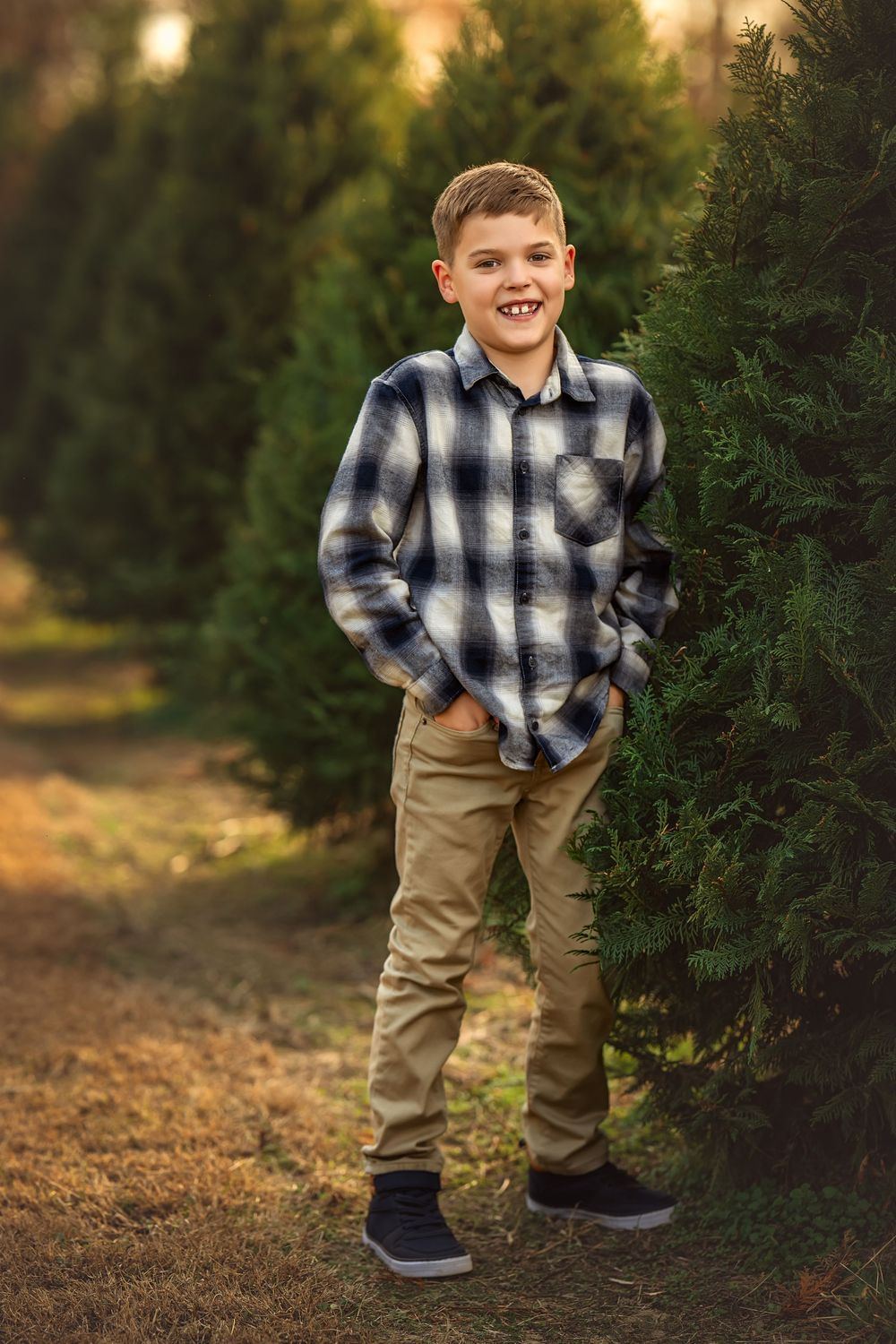Smiling boy in plaid shirt and khaki pants standing among evergreen trees at a Christmas tree farm.