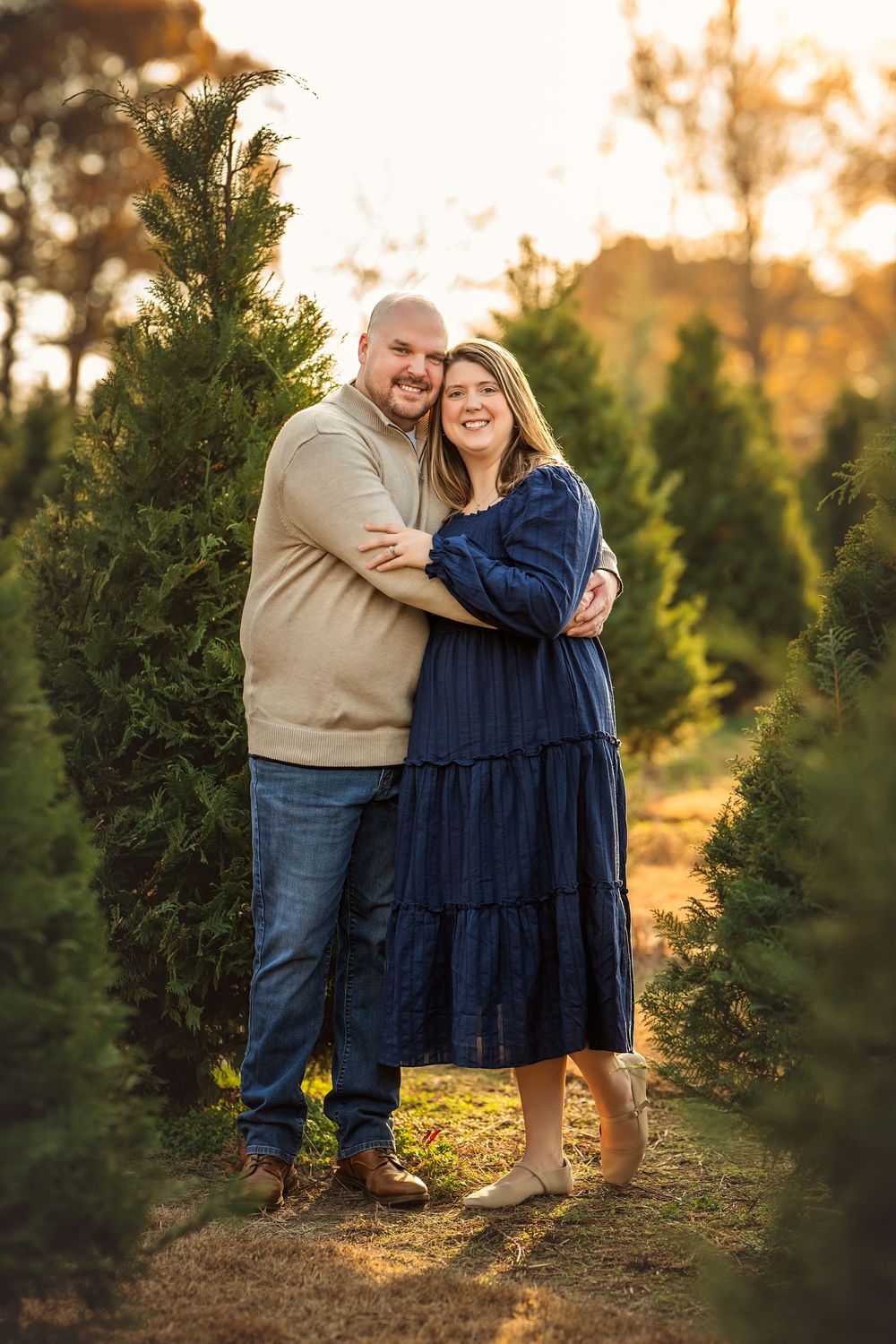 Couple embracing among evergreen trees at golden hour, woman in navy dress and man in tan sweater with jeans.