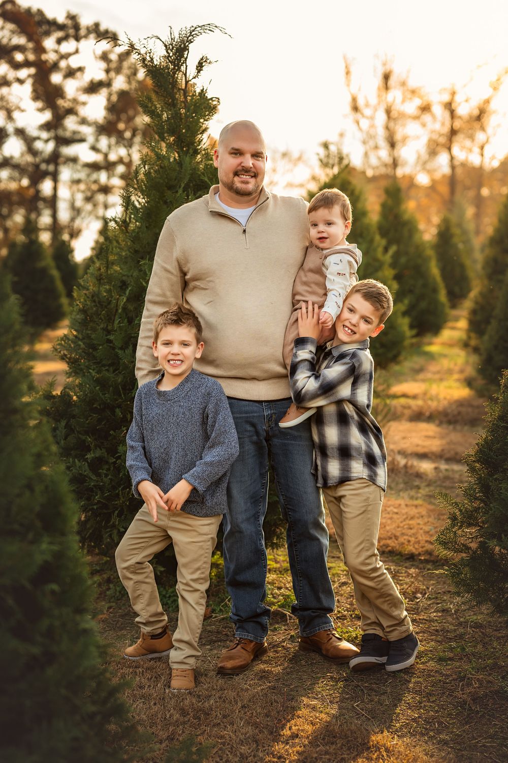 Father poses with three sons among tall evergreen trees at golden hour in a warm family Christmas tree farm portrait.