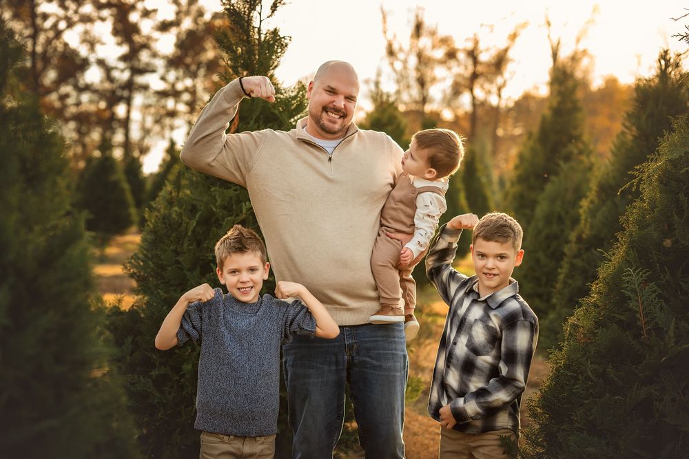 Father and three young sons pose happily among evergreen trees at a Christmas tree farm during golden hour.
