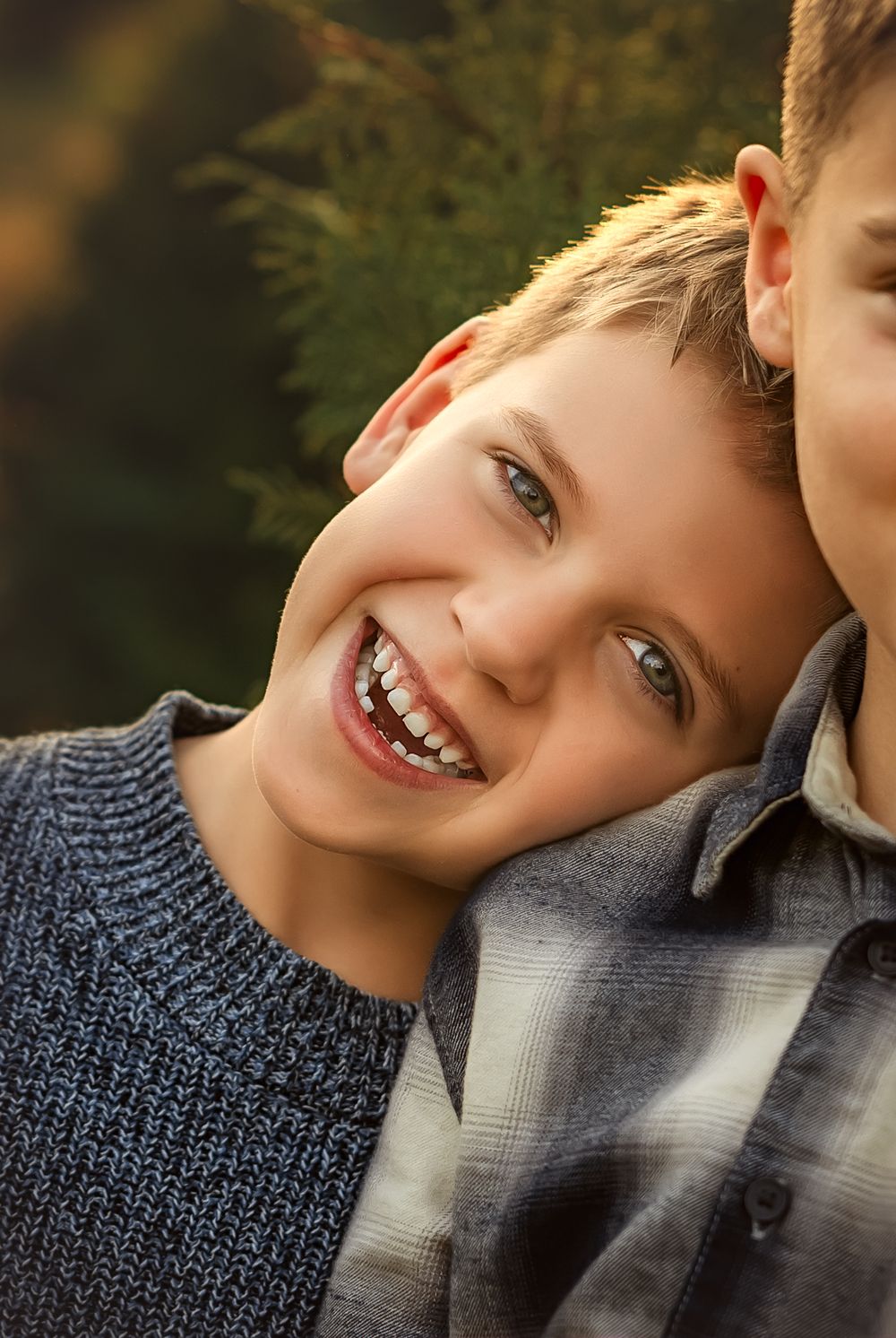 Two smiling boys lean together outdoors, sharing a joyful moment near green foliage in warm natural light.