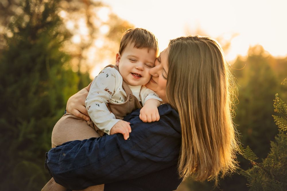A mother kisses her smiling baby boy outdoors at golden hour among green trees.
