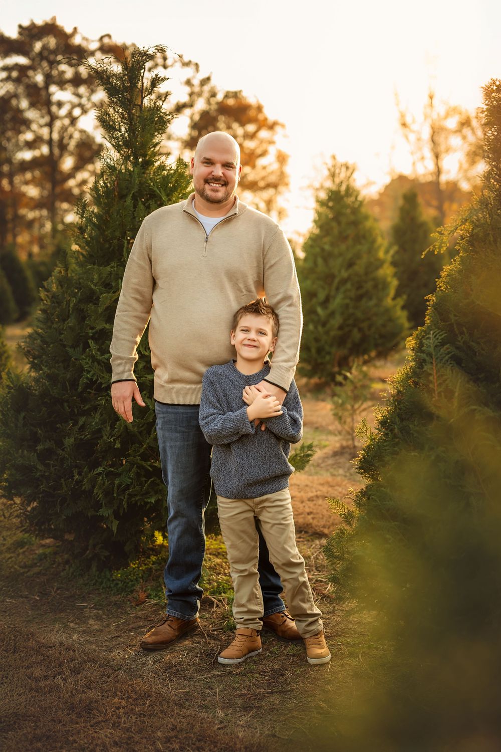 A man and young boy pose together at a Christmas tree farm during golden hour sunset.
