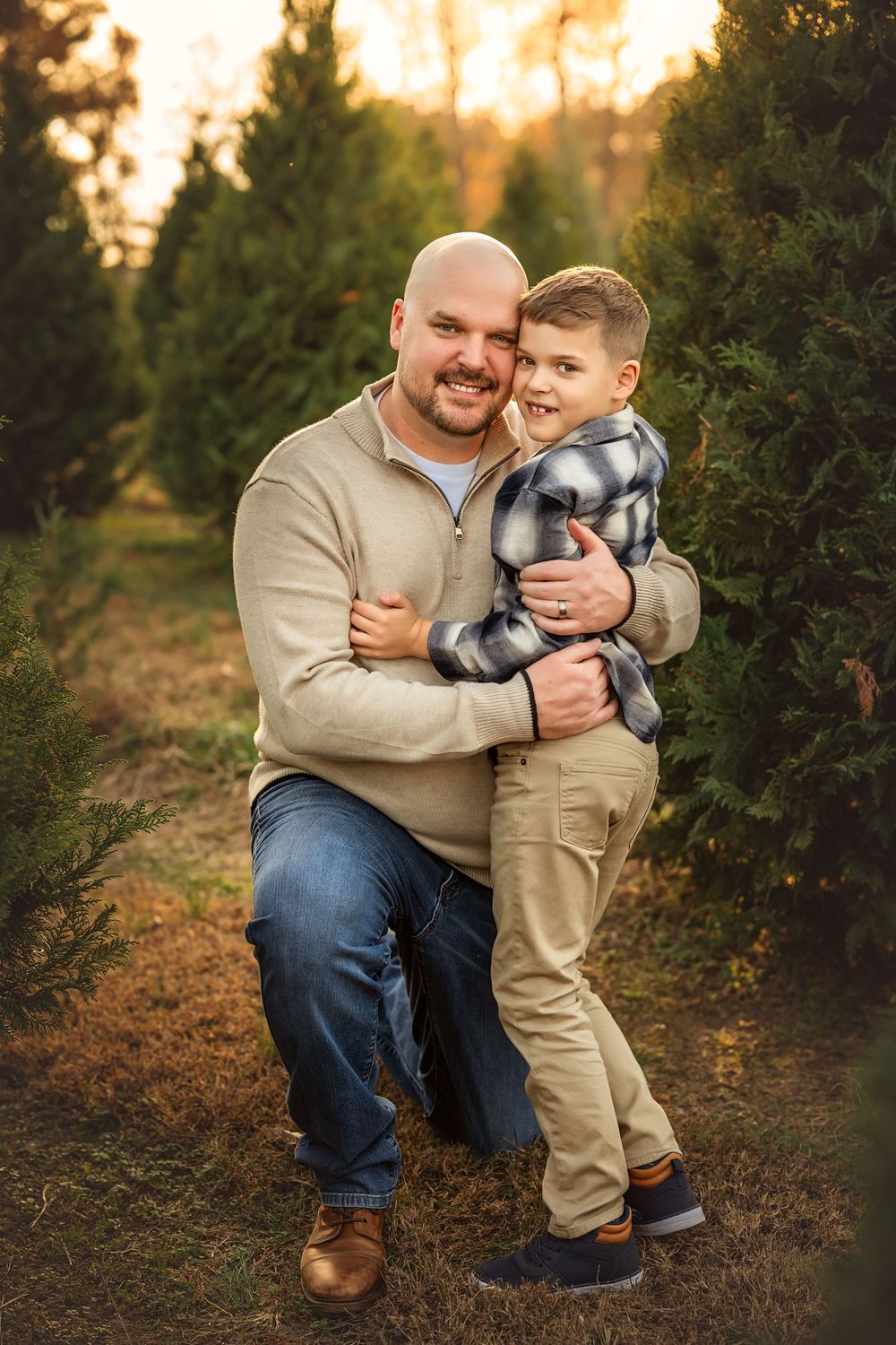 A bald father kneeling and hugging his young son at a Christmas tree farm during golden hour.
