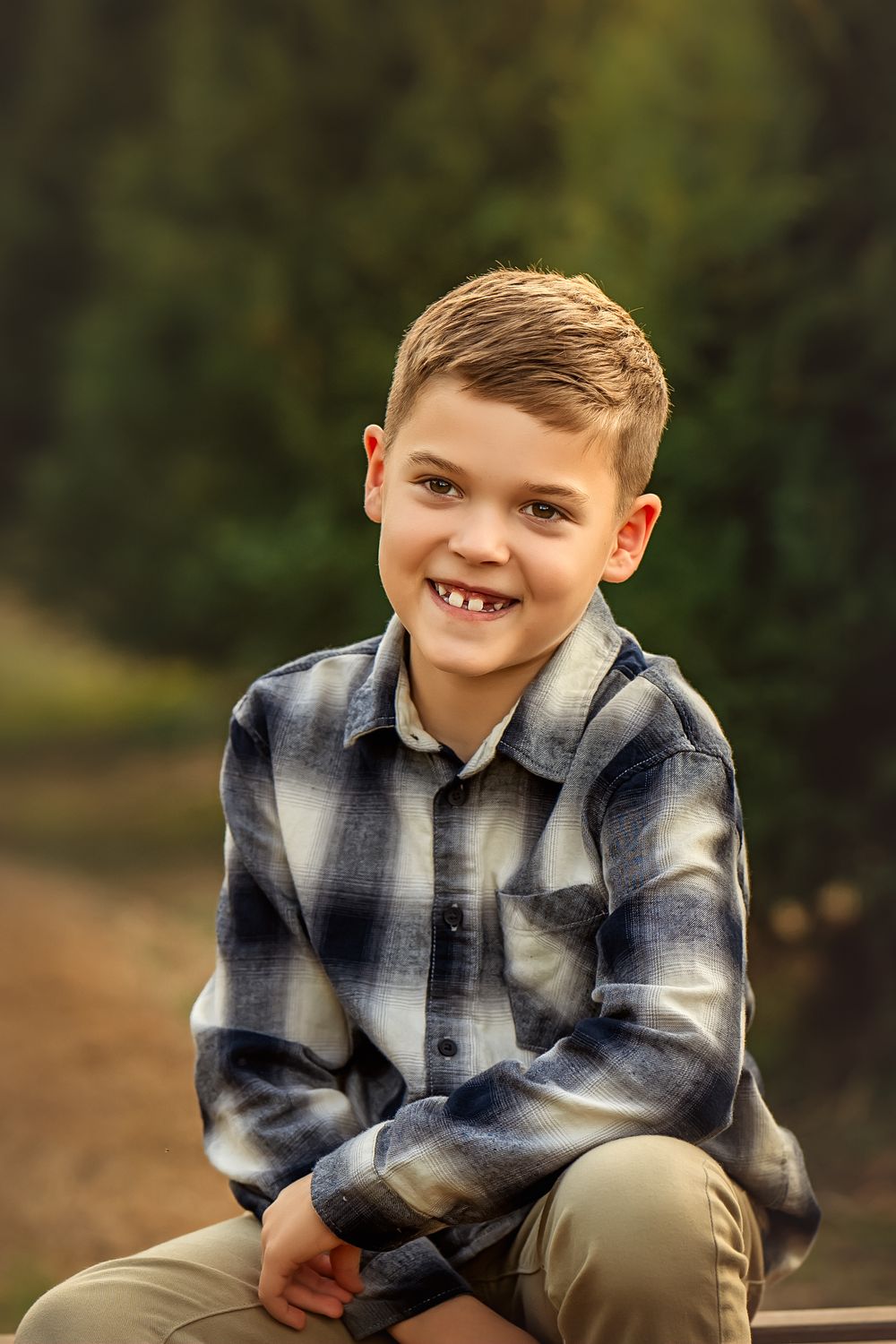 A smiling boy in a blue plaid shirt sits outdoors on a path surrounded by green trees.