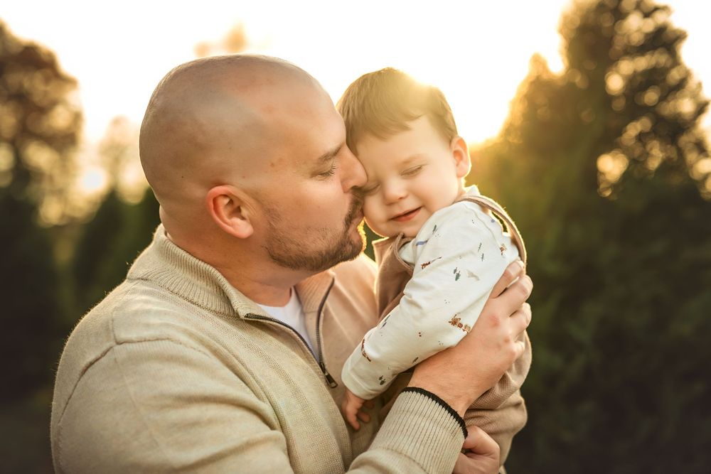A bald father lovingly kisses his smiling baby outdoors at golden hour with green trees in the background.