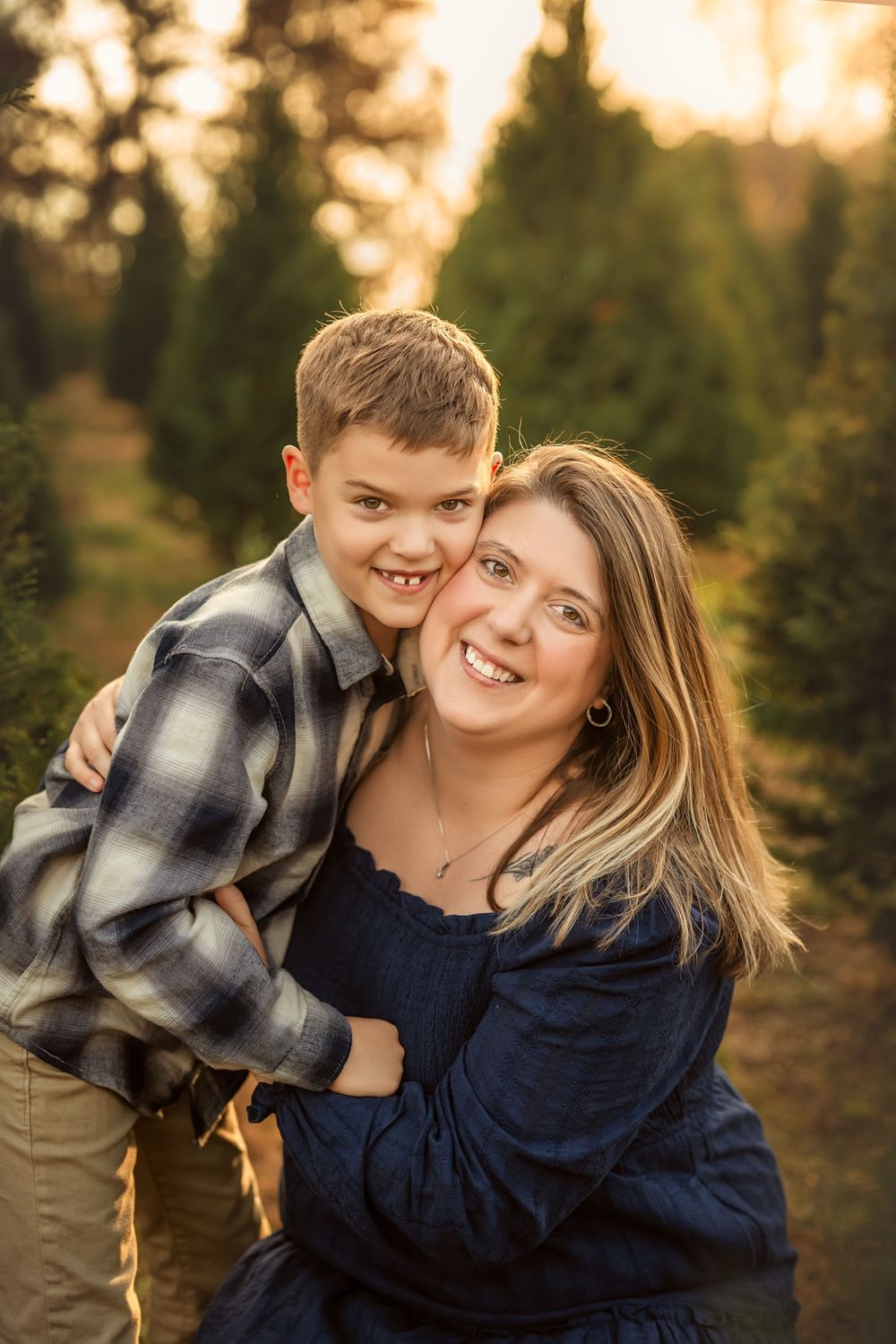 A smiling mother and young son share a warm hug at a Christmas tree farm during golden hour.