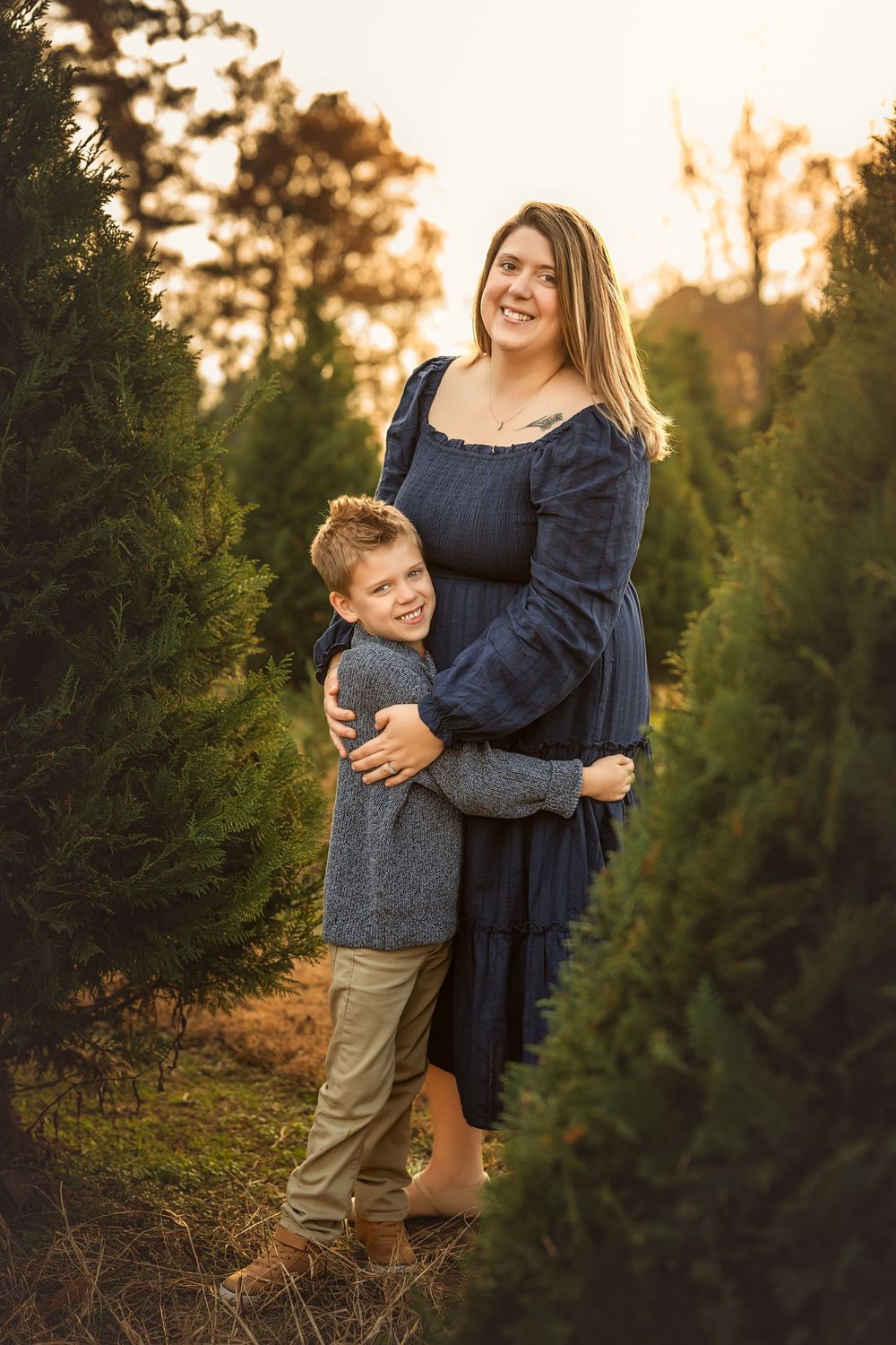 A smiling pregnant woman in a navy dress hugs her young son at a Christmas tree farm during golden hour.