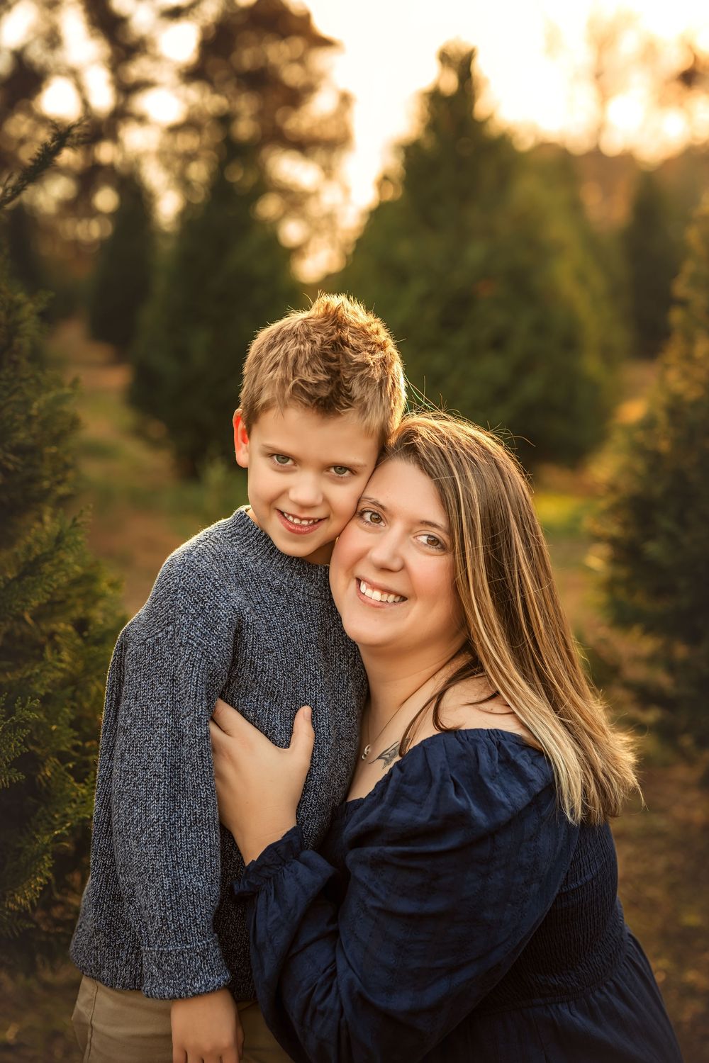 A smiling mother and young boy share a warm hug at a Christmas tree farm during golden hour.