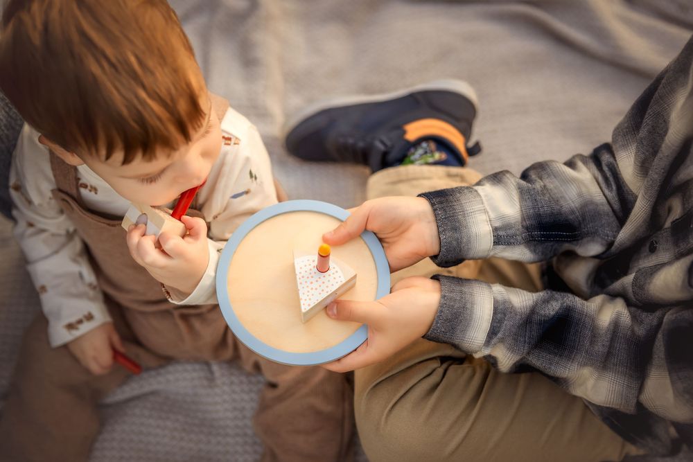 Adult feeding a toddler from a blue bowl while sitting together on a soft blanket indoors.