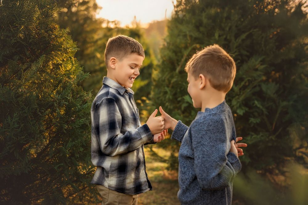 Two boys smiling and sharing a moment among green Christmas trees at a festive tree farm during golden hour.