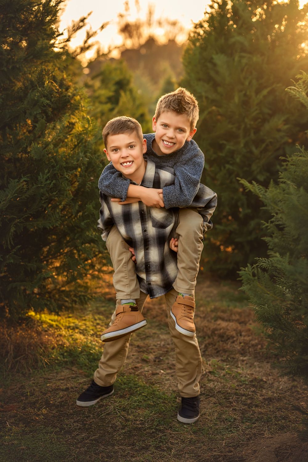 Two smiling boys enjoy a piggyback ride among evergreen trees at golden hour sunset.