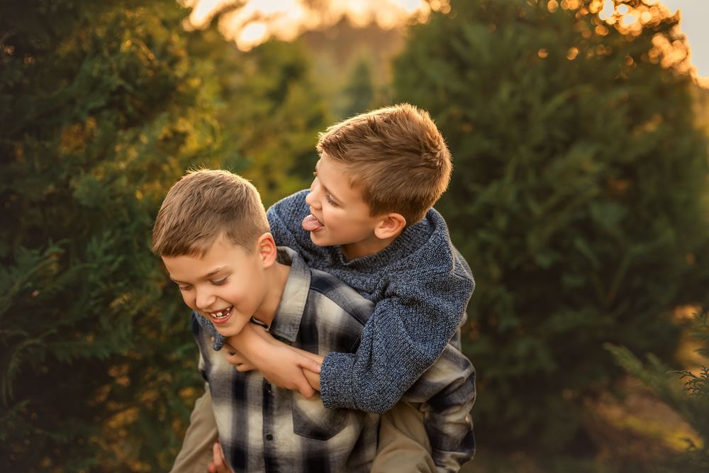 Two young brothers laugh and play piggyback at a Christmas tree farm during golden hour sunset.