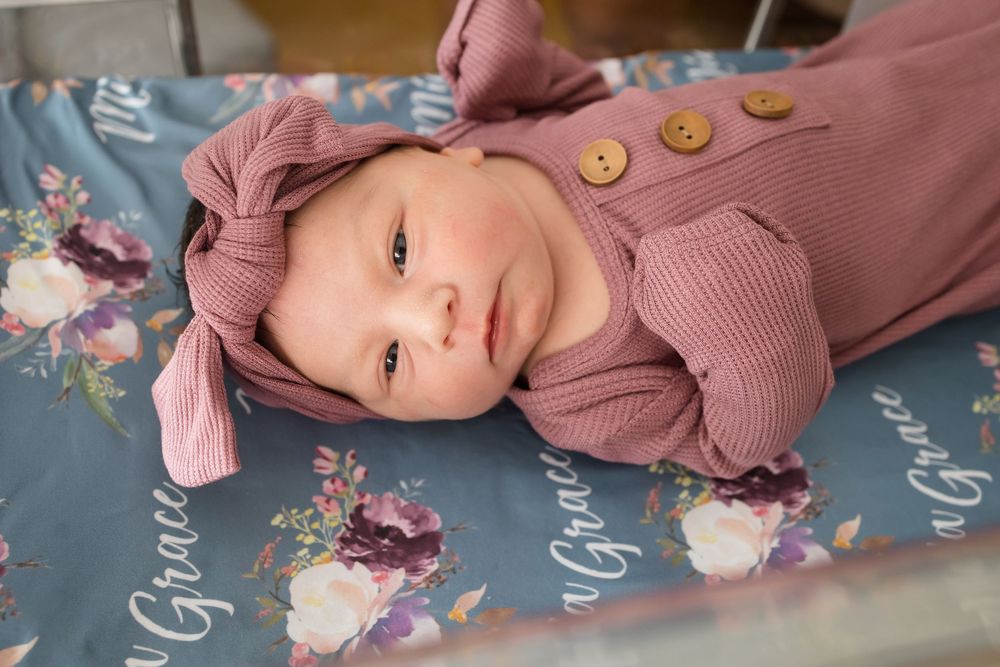 Newborn baby girl in mauve romper and bow headband lying on floral Mia Grace personalized blanket.
