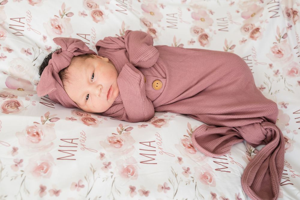 Newborn baby girl swaddled in mauve knit wrap with bow headband, lying on personalized floral blanket named Mia.