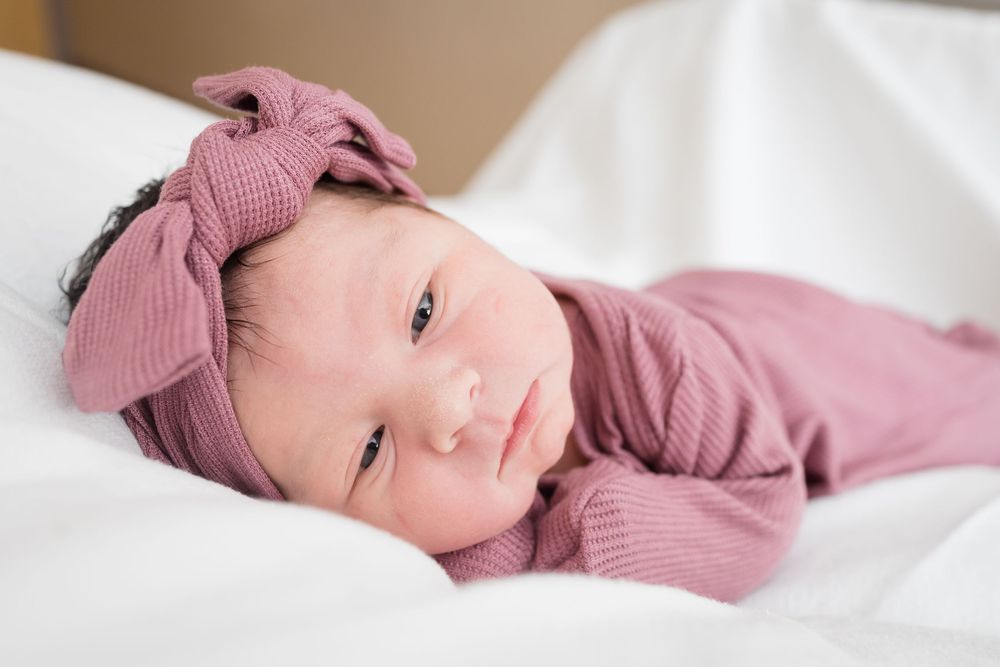 Newborn baby girl in pink outfit and bow headband lying on white blanket, wide-eyed and alert.