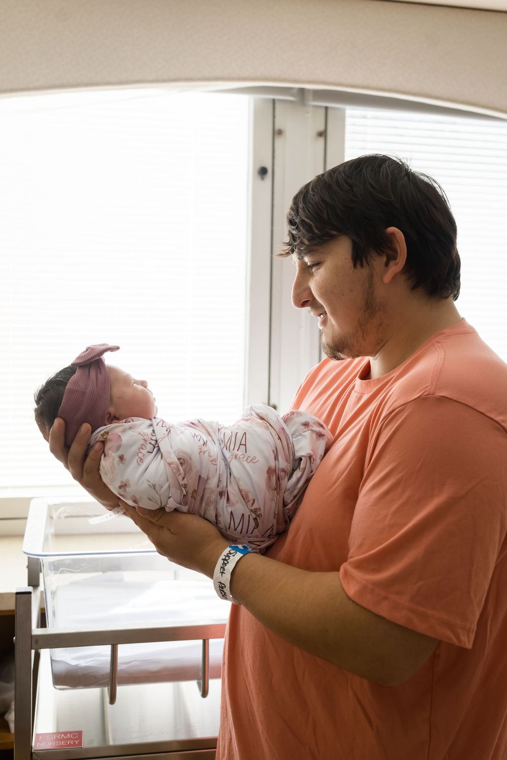 New father smiling while holding his swaddled newborn baby in a hospital room near a window.